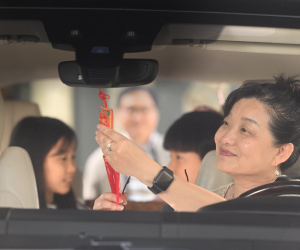 While an eldrely East Asian man loads a car, an elderly East Asian woman admires the Chinese amulet in her car while grandkids sit in the back seats talking with each other.