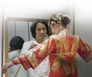 An Asian male taking measurements of an Asian female trying on a traditional Asian female wedding gown.